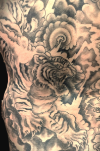Ausschnitt eines großflächigen japanischen Rückentattoos in Black-and-Grey. Zu sehen ist ein detailreich gestreifter, brüllender Tiger (Tora) inmitten von traditionellen, dunkel schattierten Sturmwolken und scharfkantigen, hellen Blitzen, gestochen von BIG WAVE TATTOO PRIVATE & ART.