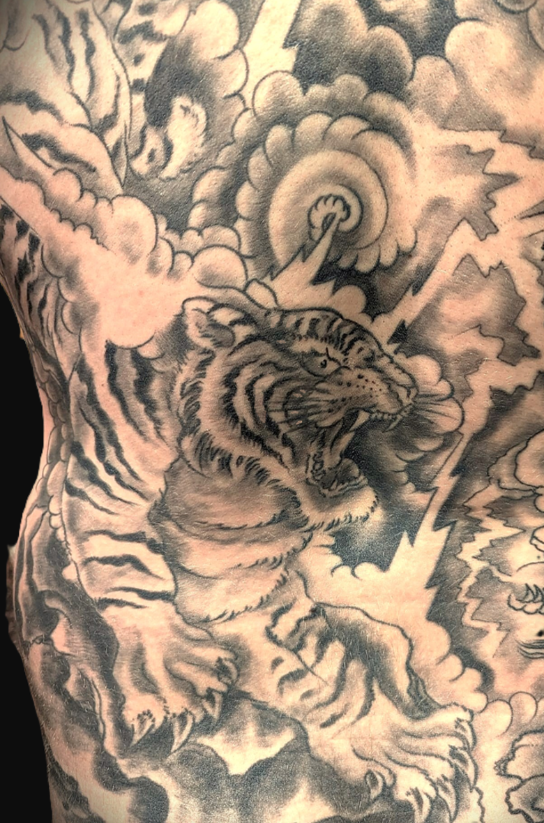 Ausschnitt eines großflächigen japanischen Rückentattoos in Black-and-Grey. Zu sehen ist ein detailreich gestreifter, brüllender Tiger (Tora) inmitten von traditionellen, dunkel schattierten Sturmwolken und scharfkantigen, hellen Blitzen, gestochen von BIG WAVE TATTOO PRIVATE & ART.
