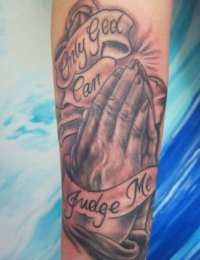 Nahaufnahme eines Unterarm-Sleeves im Black-and-Grey-Stil, das betende Hände mit strahlenden Lichteffekten im Hintergrund zeigt, umrahmt von geschwungenen Bannern mit dem Text „Only God Can Judge Me“, gestochen von BIG WAVE TATTOO PRIVATE & ART.