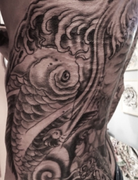 Großflächiges japanisches Tattoo auf den Rippen und der Körperseite (Rib Piece) eines Mannes. Zu sehen ist ein detailgetreuer Koi-Karpfen in feinen Black-and-Grey-Schattierungen, der sich nach oben bewegt. Der Hintergrund besteht aus traditionellen, dunklen japanischen Wellenstrukturen, Wasserstrudeln und feiner Gischt, gestochen von BIG WAVE TATTOO PRIVATE & ART.