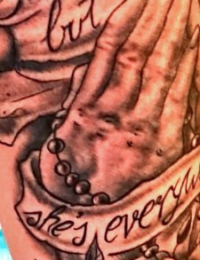 Nahaufnahme eines Arm-Tattoos im Traditional-Stil, das betende Hände mit einem Rosenkranz zeigt, umrahmt von einer großen Rose und zwei geschwungenen Schriftbannern mit dem Text „She's gone but she's everywhere“, gestochen von BIG WAVE TATTOO PRIVATE & ART.