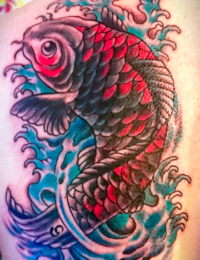 Nahaufnahme eines großflächigen japanischen Tattoos eines Koi-Karpfens auf der Schulter. Der Fisch zeigt ein Schuppenmuster in dunklen Graustufen mit leuchtend roten Farbakzenten auf dem Rücken und am Kopf. Er schwimmt in einem detailreichen Hintergrund aus dynamischen, blau und türkis schattierten Wellenstrukturen mit weißer Gischt, gestochen von BIG WAVE TATTOO PRIVATE & ART.