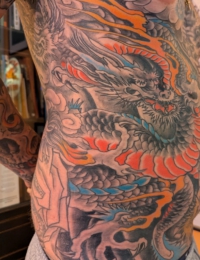 Großflächiges japanisches Tattoo auf den Rippen und dem Bauch eines Mannes. Zu sehen ist ein traditioneller Drache (Ryu) in detailreichen Black-and-Grey-Schattierungen, kombiniert mit kräftigen Akzenten in Orange, Rot und hellem Türkis. Der Hintergrund besteht aus klassischen japanischen Wolken und Flammen, gestochen von BIG WAVE TATTOO PRIVATE & ART.