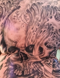 Großflächige Tätowierung auf dem gesamten Rücken eines Mannes, die einen gigantischen, detailreichen Totenkopf im Lendenbereich zeigt, aus dem dynamische, traditionell schattierte Meereswellen nach oben steigen und in eine Kette kleinerer Schädel im Nackenbereich übergehen, gestochen von BIG WAVE TATTOO PRIVATE & ART.
