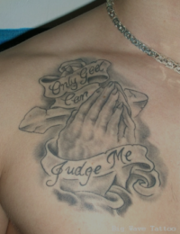 Black-and-Grey-Tattoo auf der männlichen Brust mit betenden Händen vor einem Steinkreuz, umschlossen von zwei geschwungenen Schriftbändern mit dem Schriftzug „Only God Can Judge Me“, gestochen von BIG WAVE TATTOO PRIVATE & ART in Essen.