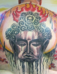 Großflächiges asiatisches Tattoo auf dem oberen Rücken. Zu sehen ist der Kopf eines meditierenden Buddhas in feinen Graustufen mit Tränen im Gesicht. Im Hintergrund steht eine große, gelb-orangefarbene Sonne, überragt von roten Flammen und flankiert von roten Kirschblüten (Sakura), gestochen von BIG WAVE TATTOO PRIVATE & ART.