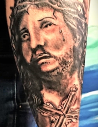 Nahaufnahme eines auf dem Unterarm tätowierten Porträts von Jesus Christus mit Dornenkrone und einer Träne im Gesicht, darunter ein verziertes Holzkreuz mit Heiligenschein im Hintergrund, gestochen von BIG WAVE TATTOO PRIVATE & ART.