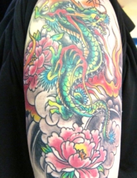 Großflächiges japanisches Tattoo auf dem Oberarm. Zu sehen ist ein detailreicher Drache (Ryu) mit grünen Schuppen, gelbem Bauch und roten Flammenausläufern. Er ist umrahmt von zwei großen pinkfarbenen Pfingstrosen (Botan) mit gelbem Kern vor einem traditionellen Hintergrund aus dunklen Wolkenstrukturen, gestochen von BIG WAVE TATTOO PRIVATE & ART.