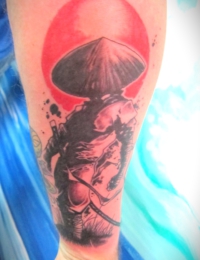 Tätowierung auf dem Unterarm im modernen japanischen Grafikstil. Zu sehen ist die Rückenansicht eines Samurai oder Rōnin mit einem traditionellen asiatischen Strohhut und einem Katana. Hinter der dunkel schattierten Silhouette steht eine große, intensiv rote Sonne. Das Motiv ist mit schwarzen Farbspritzern (Splatter-Optik) verziert, gestochen von BIG WAVE TATTOO PRIVATE & ART.