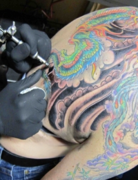 Nahaufnahme während einer Tattoo-Sitzung am oberen Rücken und Arm. Ein Tätowierer mit schwarzen Handschuhen arbeitet mit einer Maschine an einem farbintensiven japanischen Tattoo, das einen grünen Drachen, einen Phönix und eine blaue Pfingstrose vor dunklen Wolkenhintergründen zeigt, gestochen von BIG WAVE TATTOO PRIVATE & ART.