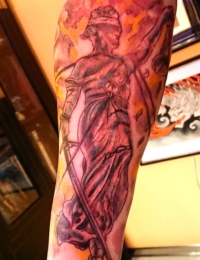 Detailliertes Black-and-Grey-Tattoo der Justitia mit Augenbinde, Richtschwert und Waage vor einem feurig schattierten Hintergrund mit dem Schriftzug „AND JUSTICE FOR ALL“ auf dem Arm, gestochen von BIG WAVE TATTOO PRIVATE & ART in Essen.