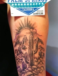 Nahaufnahme einer Arm-Tätowierung, die eine detailreiche Wüstenlandschaft mit einem großen Saguaro-Kaktus, Bergen und einer strahlenden Sonne hinter Wolken zeigt, verziert mit dezenten lila Highlights, gestochen von BIG WAVE TATTOO PRIVATE & ART.