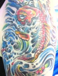 Großflächiges japanisches Oberarm-Tattoo eines Koi-Karpfens. Der detailreich geschuppte Fisch ist in kräftigen Rot-, Orange- und Gelbtönen gehalten und schwimmt aufwärts. Der Hintergrund besteht aus dynamischen blau-türkisen Wellenstrukturen mit weißer Gischt und kleinen rosa Kirschblüten (Sakura), gestochen von BIG WAVE TATTOO PRIVATE & ART.