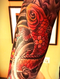 Detailansicht eines japanischen Tattoos auf dem Arm. Zu sehen ist ein kräftig rot und orange schattierter Koi-Karpfen, dessen Schuppenkleid mit kleinen rosa Kirschblüten (Sakura) mit gelbem Kern verziert ist. Der Fisch schwimmt in einem traditionellen Hintergrund aus dunklen, schattierten Wellenstrukturen, gestochen von BIG WAVE TATTOO PRIVATE & ART.