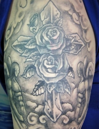 Detailansicht eines Tattoos auf dem Oberarm im Black-and-Grey-Stil. Ein steinernes Kreuz mit zwei großen Rosen steht im Mittelpunkt, flankiert von zwei geflügelten Engel-Silhouetten vor einem Hintergrund aus weichen Wolkenstrukturen. Oben und unten verläuft der geschwungene englische Schriftzug „Something to believe in“, gestochen von BIG WAVE TATTOO PRIVATE & ART.