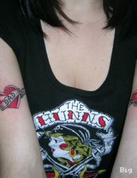 Symmetrische Tätowierungen auf den Innenseiten beider Oberarme einer Frau, die jeweils ein leuchtend rotes Herz im Old-School-Stil mit einem geschwungenen Banner und den Schriftzügen „MOM“ und „DAD“ zeigen, gestochen von BIG WAVE TATTOO PRIVATE & ART.