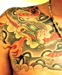 Großflächiges japanisches Tattoo auf der Brust und der rechten Schulter eines Mannes. Zu sehen ist eine detailreich geschuppte Schlange (Hebi) mit weit geöffnetem Maul, spitzen Zähnen und roter Zunge. Der rot-orange gestreifte Körper windet sich durch einen traditionellen, dicht schattierten Hintergrund aus dunklen Wolkenstrukturen, gestochen von BIG WAVE TATTOO PRIVATE & ART.