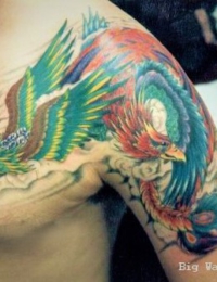 Großflächiges japanisches Tattoo auf der Brust und dem Oberarm eines Mannes. Zu sehen sind zwei detailreiche Phönixe (Hōō) mit leuchtend grünen, türkisfarbenen und roten Federn im stürmischen Flug. Der Hintergrund besteht aus klassischen, weich in Graustufen schattierten Wolkenwirbeln, gestochen von BIG WAVE TATTOO PRIVATE & ART.