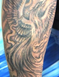 Detailansicht eines japanischen Tattoos im Black-and-Grey-Stil auf einer Extremität. Zu sehen ist ein aufwärts fliegender Phönix (Hōō) mit ausgebreiteten Flügeln und langen Schwanzfedern. Der Hintergrund besteht aus weich schattierten, wirbelnden japanischen Rauch- und Wolkenstrukturen, gestochen von BIG WAVE TATTOO PRIVATE & ART.