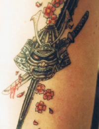 Detailreiches japanisches Tattoo eines traditionellen Samurai-Helms (Kabuto) und einer Gesichtsmaske (Mempo). Dahinter kreuzen sich zwei Katana-Schwerter, von denen eines rote Blutspritzer aufweist. Das Motiv wird von roten und rosa Kirschblüten (Sakura) mit fallenden Blütenblättern umgeben, gestochen von BIG WAVE TATTOO PRIVATE & ART.