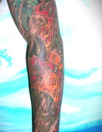 Ein vollständig tätowierter Arm im japanischen Irezumi-Stil. Auf dem Unterarm befindet sich eine traditionelle Figur im gemusterten orangefarbenen Kimono. Der obere Armbereich ist mit detailreichen roten Flammen, dunklen Schattierungen sowie grünen japanischen Ahornblättern (Momiji) gestaltet, gestochen von BIG WAVE TATTOO PRIVATE & ART.