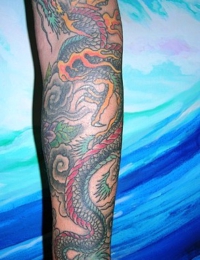 Ein vollständig tätowierter Arm (Full Sleeve) im traditionellen japanischen Irezumi-Stil. Zu sehen ist ein langgestreckter Drache (Ryu) mit dunkelgrünem Schuppenkleid und intensiv orange-magentafarbenen Ausläufern. Der Hintergrund besteht aus dichten, grau schattierten Sturmwolken und Windhosen, gestochen von BIG WAVE TATTOO PRIVATE & ART.