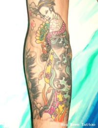 Detailreiches japanisches Tattoo einer Geisha auf dem Unterarm. Die Figur trägt einen gemusterten Kimono mit Farbakzenten in Gelb, Grün und Magenta sowie eine traditionelle Hochsteckfrisur. Der Hintergrund ist mit klassischen, dunkel schattierten Wellen- und Wolkenstrukturen gestaltet, gestochen von BIG WAVE TATTOO PRIVATE & ART.