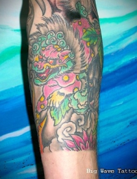 Detailansicht eines bunten japanischen Arm-Tattoos. Zu sehen ist ein Wächterlöwe (Karashishi) mit pinkfarbenem Körper, gelben Mustern und grüner Mähne, der sich an grauen Felsen hinabbewegt. Am unteren Abschluss befindet sich eine pinke Lotusblüte vor dunklen Wolkenstrukturen, gestochen von BIG WAVE TATTOO PRIVATE & ART.