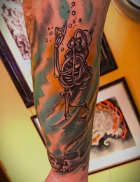 Traditional Tattoo eines Skelett-Tauchers in historischer Taucherausrüstung, umgeben von Luftblasen vor einem wellenartigen Hintergrund in Grün- und Orangetönen, gestochen von BIG WAVE TATTOO PRIVATE & ART in Essen.