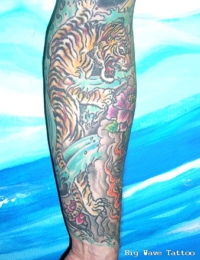 Detailansicht eines farbigen japanischen Arm-Tattoos (Sleeve). Zu sehen ist ein orange-gelb gestreifter Tiger (Tora) mit geöffnetem Maul, der an einem schattierten Felsen hinabklettert. Rechts daneben befindet sich eine pinke Pfingstrose, eingebettet in wirbelnde blau-türkise Wellenstrukturen, gestochen von BIG WAVE TATTOO PRIVATE & ART.