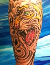 Detailansicht eines japanischen Tattoos auf einer Extremität. Zu sehen ist der Kopf eines zornig brüllenden Tigers (Tora) mit geöffnetem Maul, scharfen Zähnen und funkelnden grünen Augen. Das Fell ist in Orange-Gelb gestochen und von dichten, traditionell schwarzen Wolkenwirbeln umgeben, gefertigt von BIG WAVE TATTOO PRIVATE & ART.