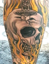 Nahaufnahme eines auf dem Unterschenkel tätowierten Totenkopfs im Traditional-Stil, der von oben nach unten von einem verzierten Dolch durchstochen wird und von gelb-orangen Flammen umgeben ist, gestochen von BIG WAVE TATTOO PRIVATE & ART.