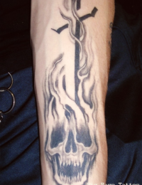 Nahaufnahme eines auf dem Unterarm tätowierten Schädels im Traditional-Stil, aus dem dichte Flammen und Rauch aufsteigen, die ein großes Kreuz sowie fliegende Vögel umhüllen, gestochen von BIG WAVE TATTOO PRIVATE & ART.