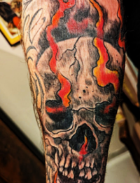 Detailansicht eines Unterarm-Tattoos im traditionellen Stil. Zu sehen ist ein detailreich schattierter Totenkopf in Black-and-Grey, durch dessen Risse, Nase und Augenhöhlen intensiv rote und orangefarbene Flammen nach oben schlagen. Die Flammen bilden auch den unteren Abschluss am Handgelenk, gestochen von BIG WAVE TATTOO PRIVATE & ART.