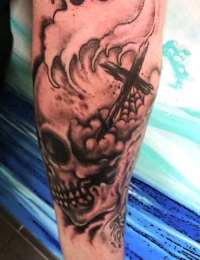 Nahaufnahme eines auf dem Unterarm tätowierten Totenkopfs im Traditional-Stil, der mit einem hölzernen Kreuz, einem Spinnennetz in der Augenhöhle und dynamischen Wellen verziert ist, gestochen von BIG WAVE TATTOO PRIVATE & ART.