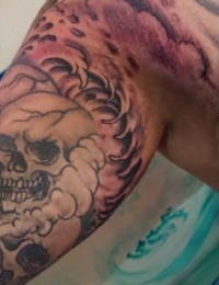 Nahaufnahme eines auf dem Oberarm tätowierten Totenkopfs im Traditional-Stil, eingebettet in dynamische Wellenstrukturen und dunkle Wolken-Schattierungen, gestochen von BIG WAVE TATTOO PRIVATE & ART.