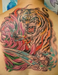 Großflächiges japanisches Tattoo auf dem oberen Rücken. Zu sehen ist ein detailreicher Tiger (Tora) mit grün leuchtenden Augen und geöffnetem Maul. Links daneben befindet sich eine große, pinkfarbene Chrysantheme mit grünen Blättern und einem Schmetterling. Im unteren Bereich fließen traditionelle blaue Wellen und Flammenstrukturen zusammen, gestochen von BIG WAVE TATTOO PRIVATE & ART.