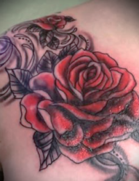 Detailansicht eines Tattoos auf dem oberen Rücken und dem Schulterblatt. Zu sehen ist eine große, leuchtend rote Rose mit schattierten Blättern. Der Hintergrund ist mit geschwungenen, schwarzen Ornament-Verzierungen und einer kleineren Rosenknospe gestaltet, gestochen von BIG WAVE TATTOO PRIVATE & ART.